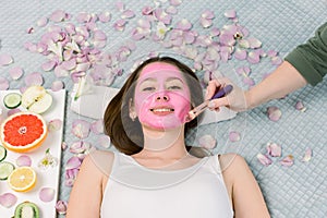 Face mask, skin care, spa procedures. Beautiful young girl at spa, lying on bed with flower petals and fresh fruits