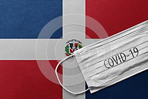 Face Medical Surgical White Mask with COVID-19 inscription lying on Dominican Republic Flag. Coronavirus in Dominicana