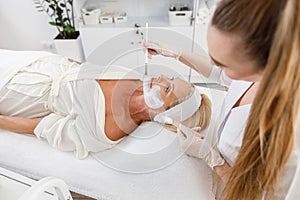 Face peeling mask, spa beauty treatment, skincare. Senior woman getting facial care by beautician at spa salon