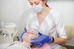 Face peeling and treatment at the beautician parlor. Woman cosmetologist applies a foam to the clients face with a hands