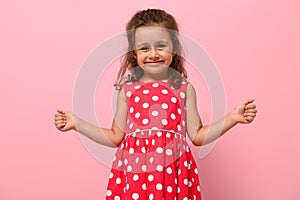 Face portrait of a beautiful child girl in pink polka dots dress, cute smiling looking at camera, posing with thumbs up, over pink