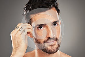 Face, portrait and tweezers with a man model plucking his eyebrows in studio on a gray background. Skincare, wellness