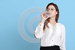 Face portrait of young attractive woman dresses white blouse and black skirt, holds glass in hand and drinks water. Dark haired