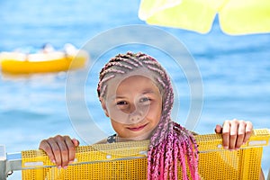 Face of pre-teen Caucasian girl with pink dreadlocks hairstyle looking out from yellow sun lounger, sunny coastline