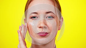 Face of slim freckled redhead young woman with green eyes looking at camera applying skin moisturizer. Close-up portrait