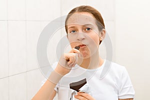 Face of a teenage girl with pimples, acne on the skin, she looks at herself in the mirror and eating a chocolate, concept of