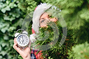 Face of woman in a santa claus red hat holding alarm clock in green fir tree branches in the forest. Christmas gift concept.