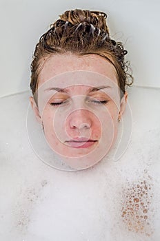 Face of a young beautiful girl in a white bath among soap bubbles from the foam bath gel, naked with wet hair, enjoys the spa trea