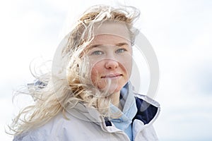 The face of a young girl with hair flying in the wind against the blue sky on a sunny day. Smiling blonde teenager in a white