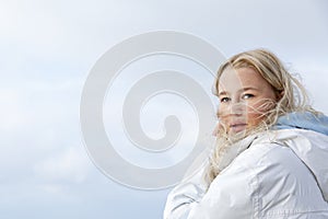 The face of a young girl with hair flying in the wind against the blue sky on a sunny day. Smiling blonde teenager in a white