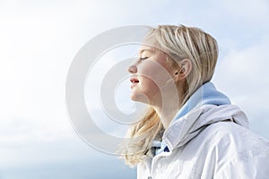 The face of a young girl with hair flying in the wind against the blue sky on a sunny day. Smiling blonde teenager in a white