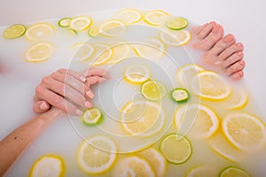 Faceless beautiful young woman takes a bath with milk lemons and lime. Cropped photo. Close-up of female hands and feet