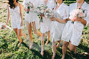 Faceless bridesmaids and bride in satin robes with bouquets in hands walk barefoot on the grass photo