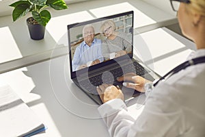 Faceless doctor sitting in her office and talking to a senior couple via video call using a laptop.