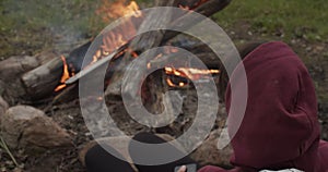 Faceless Man In Hood Sitting By The Fire Outdoors Close-up. Unrecognizable Person Warming Near Bonfire Or Spending Time