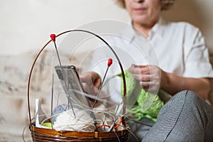 Faceless old woman knits and watches online training on a smartphone. Close-up of female hands with yarn and knitting