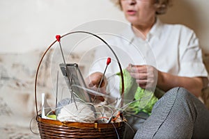 Faceless old woman knits and watches online training on a smartphone. Close-up of female hands with yarn and knitting