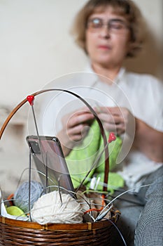 Faceless old woman knits and watches online training on a smartphone. Close-up of female hands with yarn and knitting