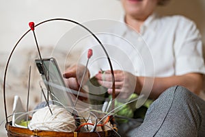 Faceless old woman knits and watches online training on a smartphone. Close-up of female hands with yarn and knitting