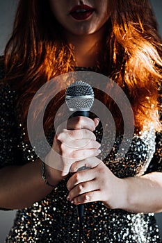 Faceless portrait of redhead woman in sparkly evening dress holding microphone on dark night background. Unrecognizable