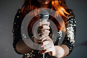 Faceless portrait of redhead woman in sparkly evening dress holding microphone on dark night background. Unrecognizable