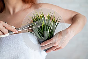 Faceless red-haired Caucasian woman wrapped in a white terry towel shears a potted plant with scissors. Armpit hair