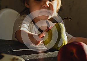 Faceless toddler, a little boy or girl sits at the table and reaches for the pear. Matte shadows, sunset color