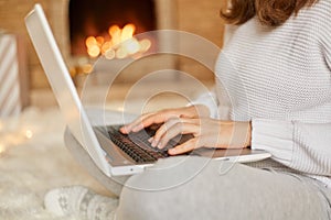 Faceless woman typing message on her laptop computer as she relaxes near burning warm fire, unknown female working online while
