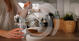 Faceless young woman pour pure water from jug to glass in kitchen. Anonymous