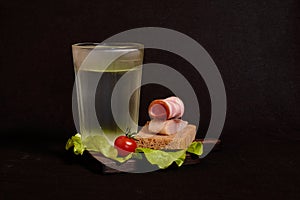 Faceted glass of vodka, sandwich with rye bread, herring, ham, tomato, lettuce