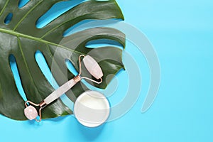 Massage roller and face cream on a monstera leaf, blue background, top view. Flat lay