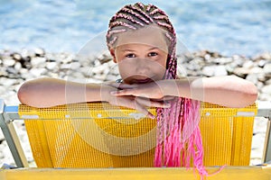 Facial portrait of pre-teen Caucasian girl with pink dreadlocks hairstyle looking out from yellow sun lounger, sunny coastline