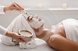 Facial treatment. Beautician applying clay face mask to asian woman at spa