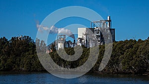 Factory building set on riverbank with steam evaporating to blue sky from chimney