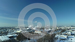 Factory buildings covered with snow. Factory smoke chimneys. There is a railway nearby. You can see the city blocks. Winter