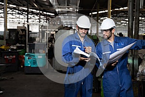 Factory engineer reading manual of machine operation and standing with blue working suite dress and safety helmet.