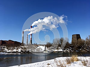 Factory pipes with white smoke near the river in winter
