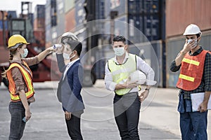 Factory woman worker in a face medical mask and safety dress used measures temperature at worker people standing on queue with a