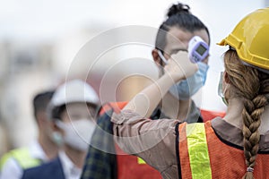 Factory woman worker in a face medical mask and safety dress used measures temperature at worker people standing on queue with a