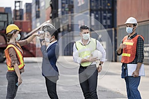 Factory woman worker in a face medical mask and safety dress used measures temperature at worker people standing on queue with a