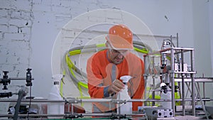 Factory worker man in helmet and workwear checks quality production of household chemicals on automatic belt line