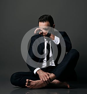 Failed adult man, bankrupt in black office suit sits barefooted on floor with legs crossed, closing face with hand