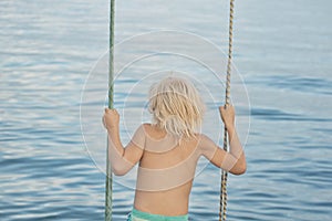 Fair-haired boy rides rope swing on sea background. Carelessness. Happy childhood photo