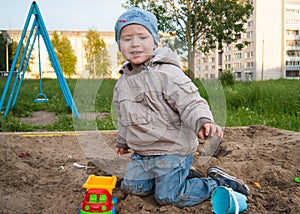 A fair-haired boy of three years in a blue hat and jeans, in a light jacket plays in the sandbox with toys