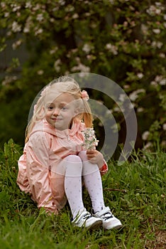 Fair-haired girl in pink dress sitting near blooming tree and looking in the camera. Cute child girl in the flowering garden.