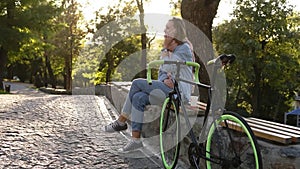 Fair haired girl sitting on the bench or parapet in the city park with paved her trekking bike next to her. Talking by