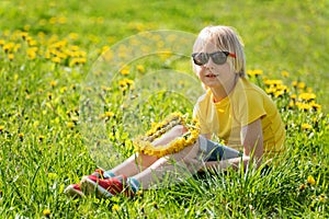 Fair-haired little child boy in yellow t-shirt sits on green grass lawn. Kid weaves wreath of dandelions sitting in field