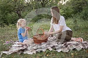 Fair-haired little girl eats apple sitting on plaid blanket in woods. Mother and daughter with wicker basket at picnic in park