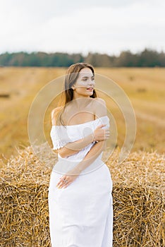 A fair-haired young woman in a white dress in a field with stacks of straw is enjoying a summer day