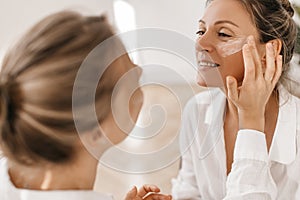 Fair-skinned girl applies moisturizer to face of adult woman on white background.